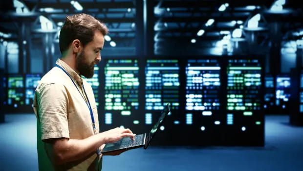 IT professional working on a laptop in a high-tech data center environment. Represents real-time monitoring and management of secure server infrastructure.