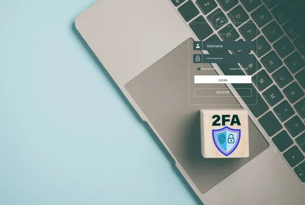 Open laptop keyboard and trackpad with a transparent login form overlay showing username and password fields. Wooden cube labeled “2FA” with a security shield icon, symbolizing two-factor authentication and account protection.